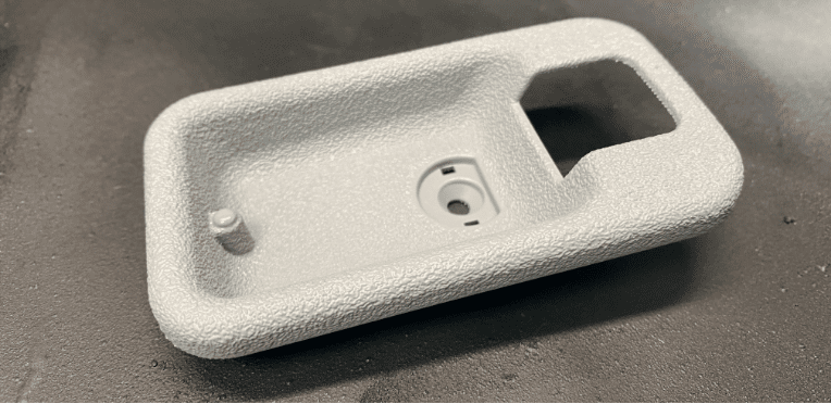 A textured, rectangular grey plastic component with a recessed section, a small raised peg, a central circular opening, and a larger cut-out at one end, placed on a dark surface.