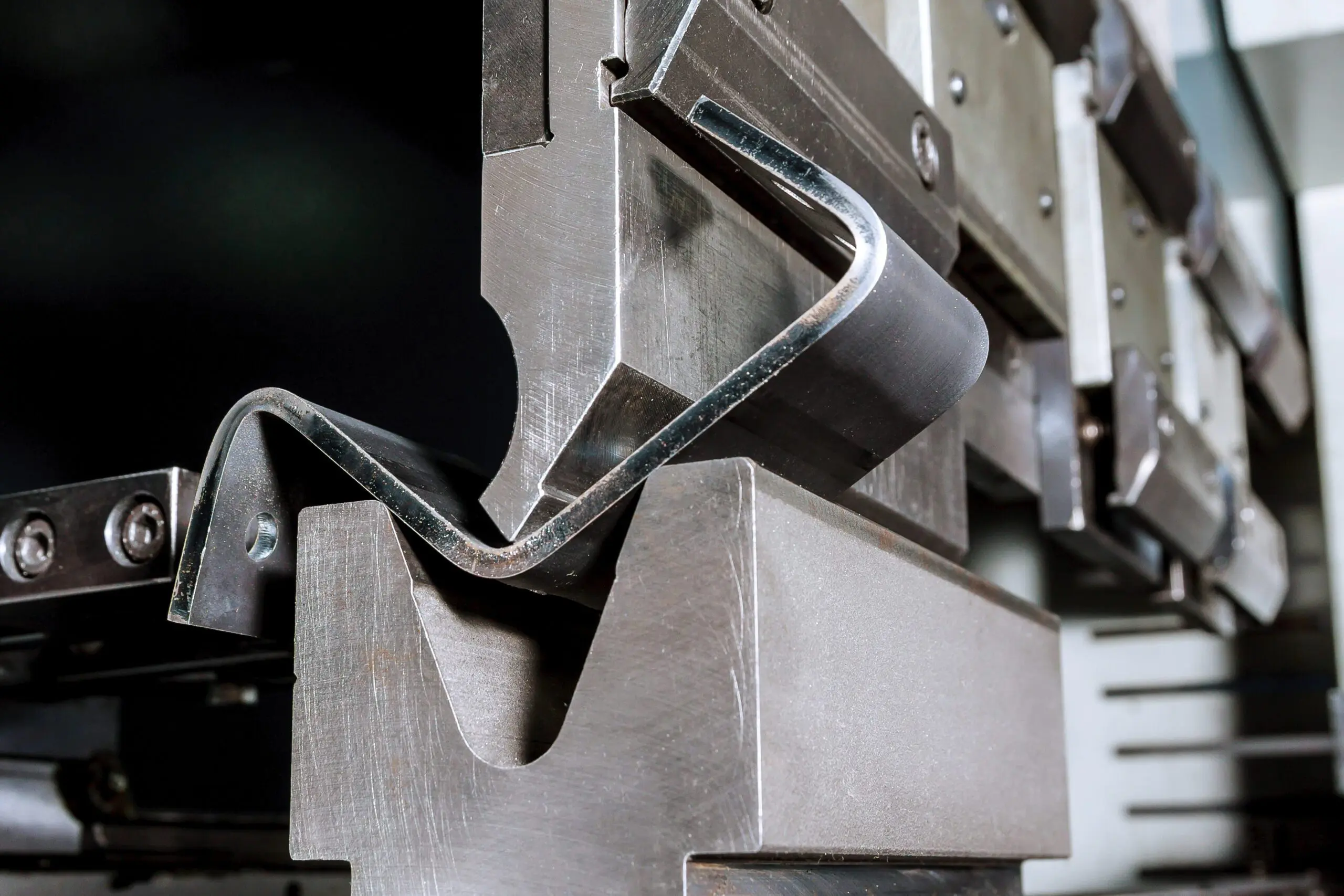 Close-up of a press brake machine bending a metal sheet to a precise angle, showing the tooling and partially formed metal component.