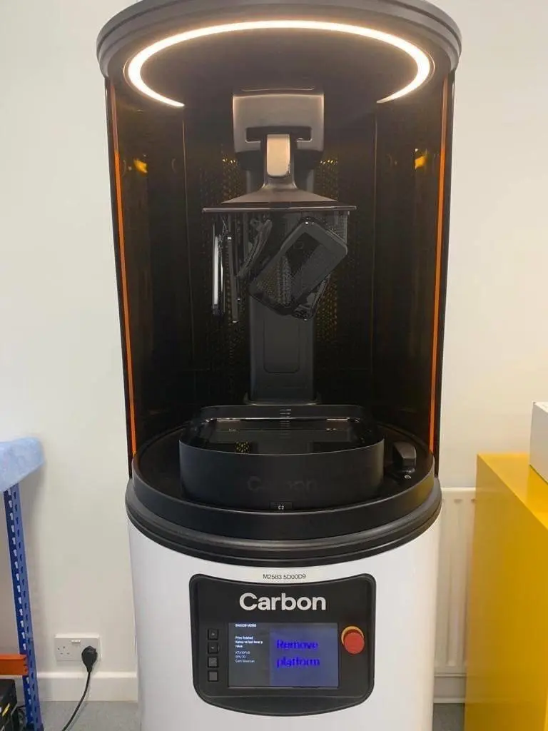 A Carbon 3D printer with a black object suspended from the build platform, lit by a circular LED light, with a visible control panel and emergency stop button.