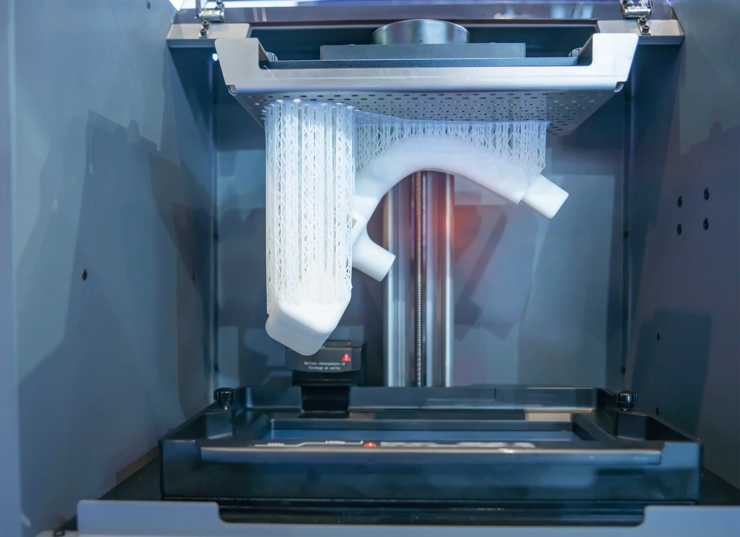 A 3D printer producing a detailed white plastic object with intricate lattice support structures inside the build chamber.