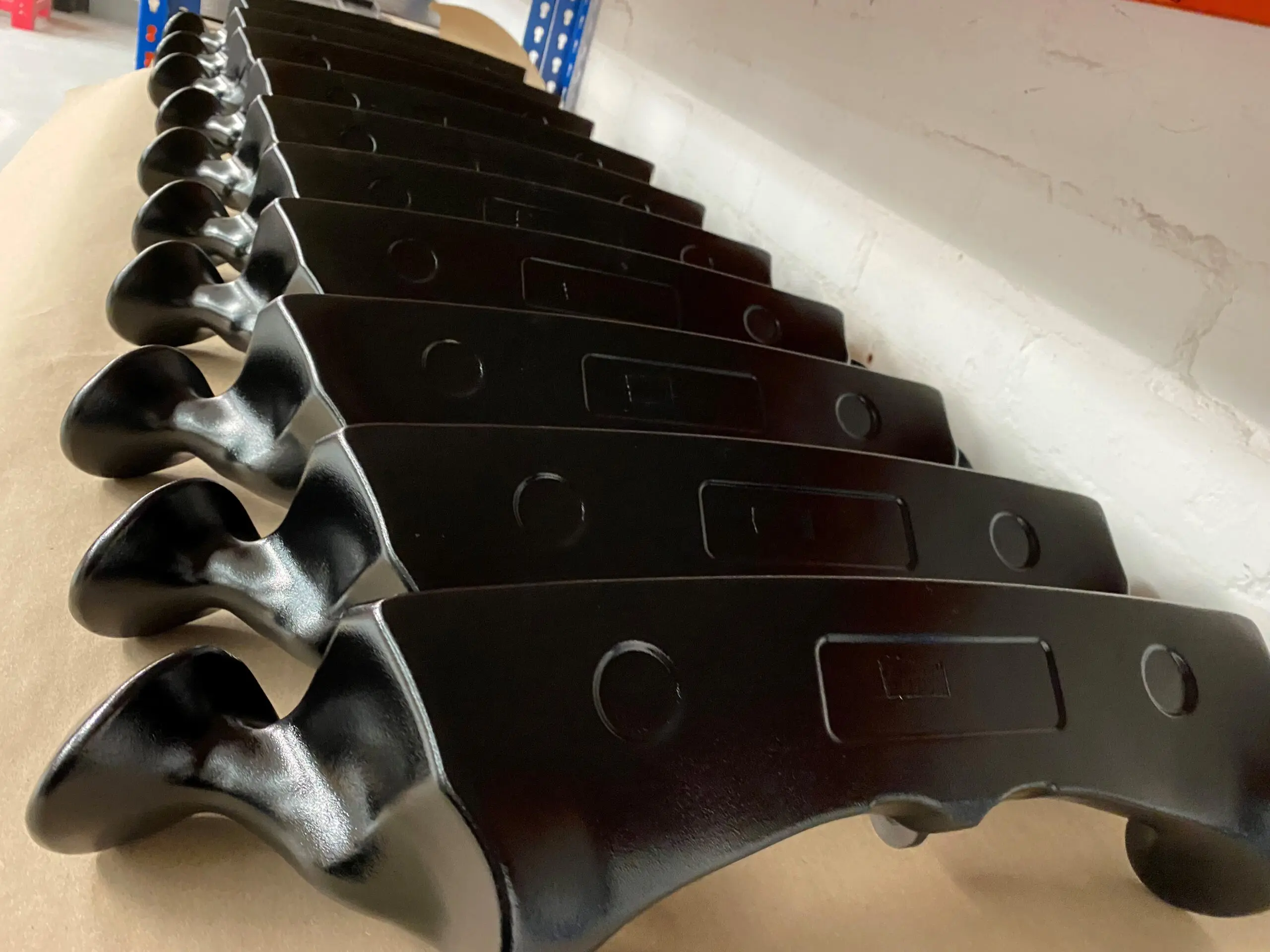 A row of identical black metal handles or brackets lined up on a sheet of brown paper, placed on a workbench with a white brick wall in the background.