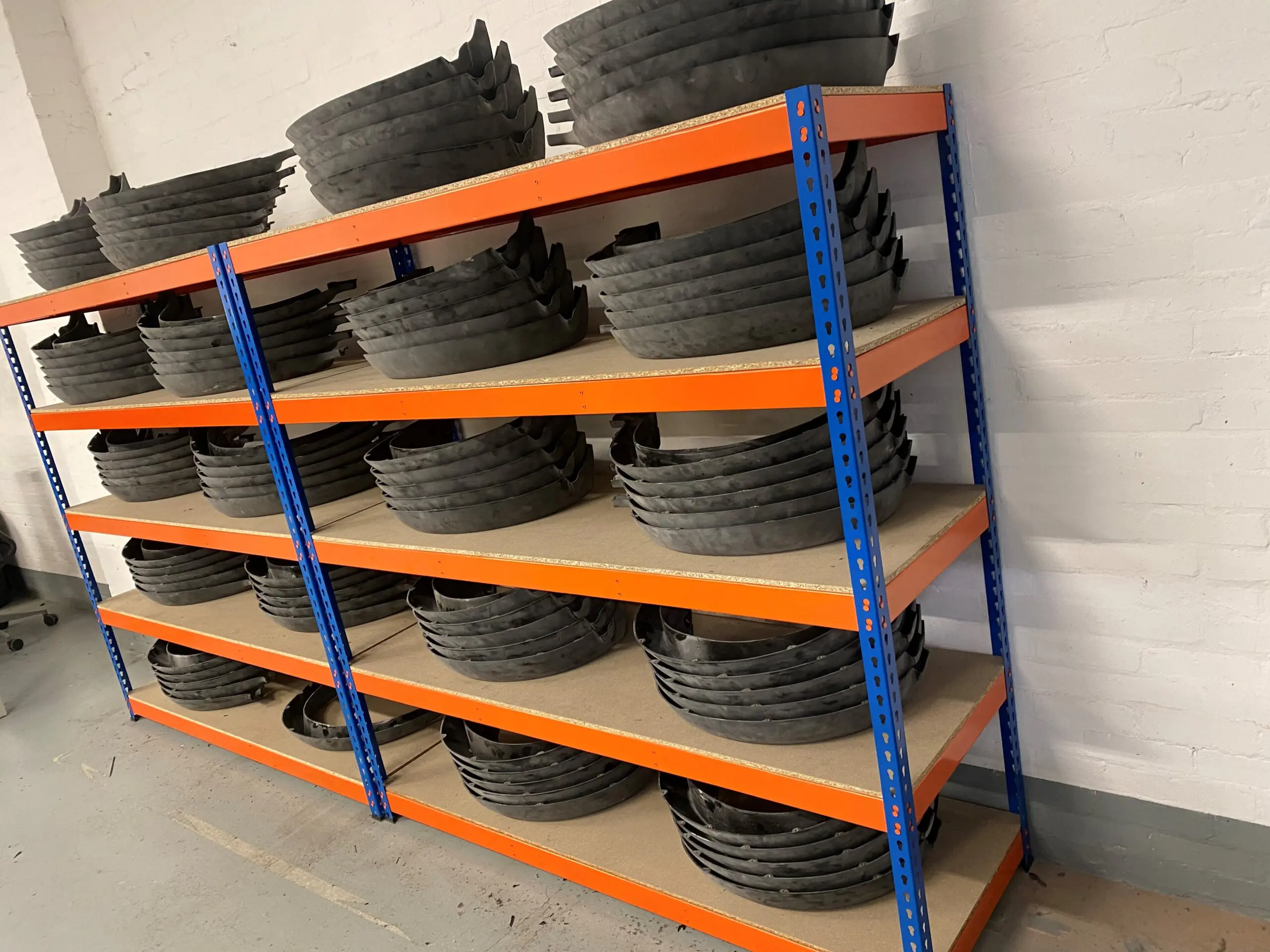 Metal shelves with orange beams and blue supports hold multiple stacks of black, curved plastic or rubber pieces, all neatly organised. The shelving unit is set against a white brick wall on a grey floor.