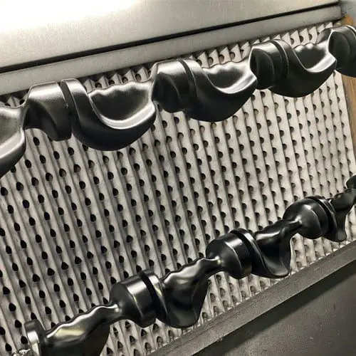 Two black camshafts with intricate lobes displayed against a metallic perforated background, showing machined surfaces and complex shapes.