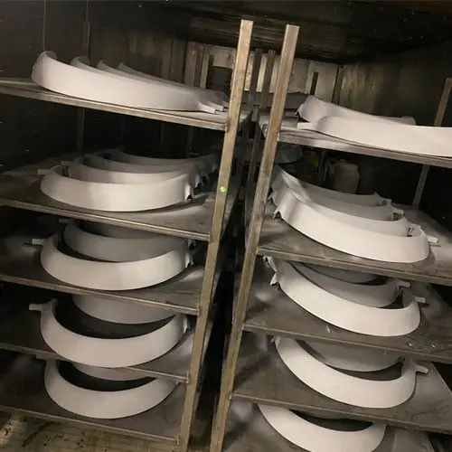 Stacks of curved white plastic or fibreglass pieces neatly arranged on metal shelves inside an industrial oven or drying rack.